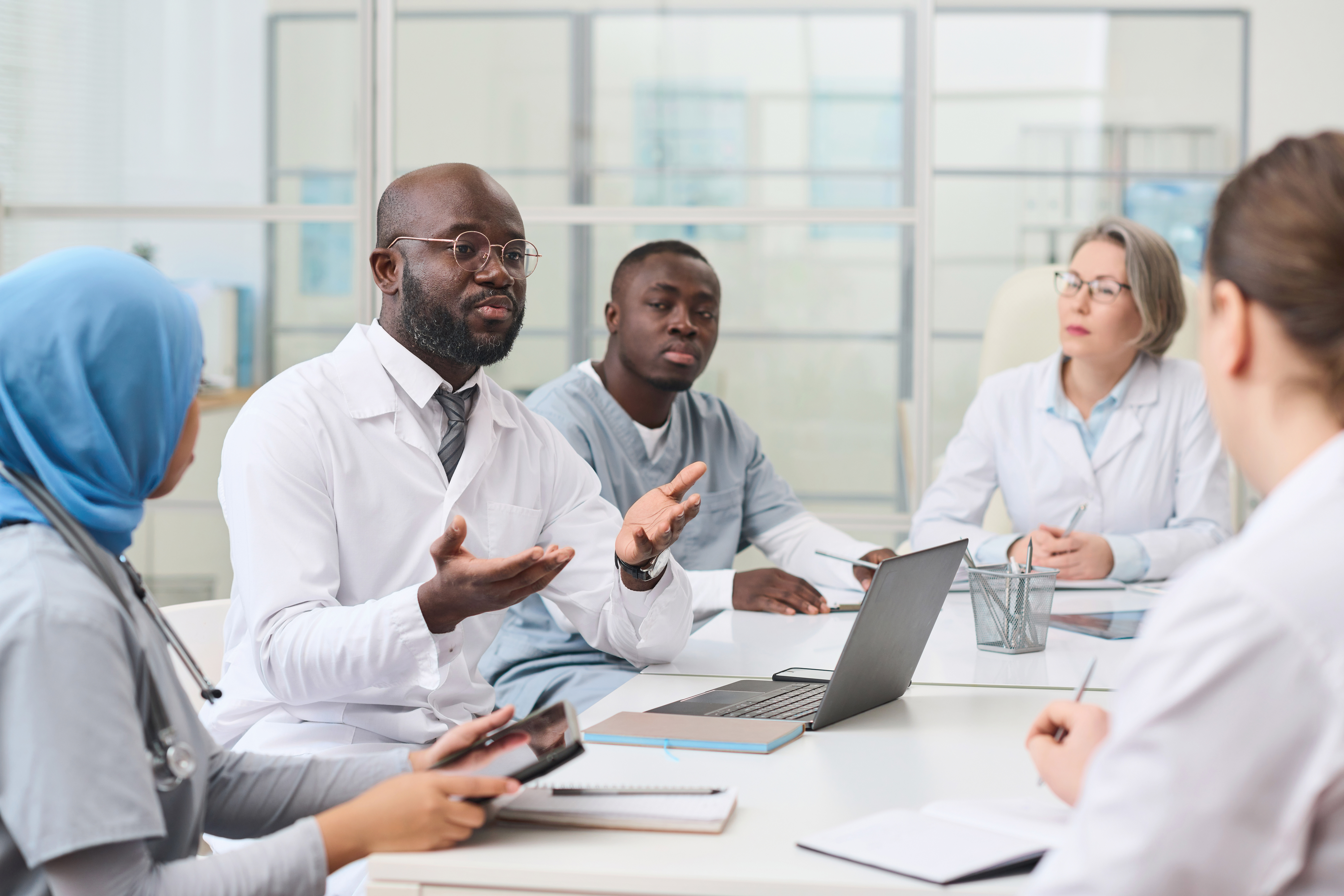 A diverse group of healthcare professionals sit around a conference table engaged in a discussion, with one doctor actively speaking while others listen and take notes.